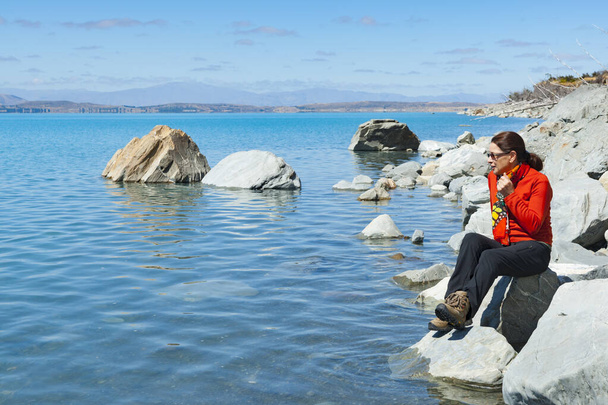 Nainen turisti oranssi alkuun istuu kiviä järven reunalla ja turkoosi sininen vesi lumen rehun luonnonkaunis järvi Pukaiki Etelä-Ilsand Uusi-Seelanti. - Valokuva, kuva