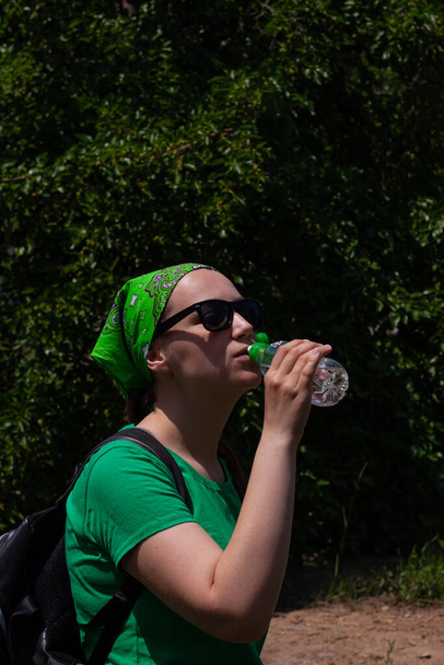 Mladá dívka drží v ruce láhev vody a pije. Portrét pěkného hipstera ve slunečních brýlích, zelené košili, šátek stojí na přírodě za slunečného dne. Hubnutí, letní aktivní životní styl péče o tělo. - Fotografie, Obrázek