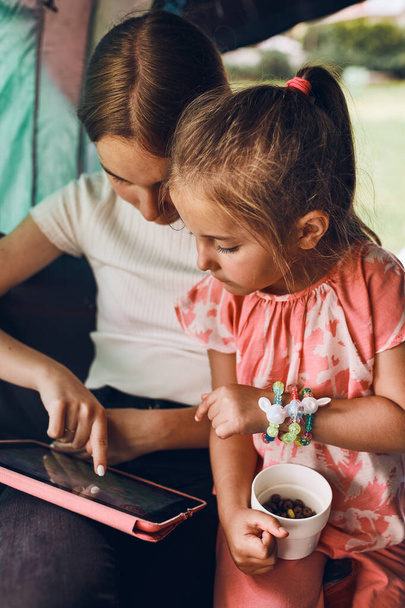 Two girls sisters spending family time in a tent on camping. Children using tablet playing games online during summer vacation - Foto, afbeelding