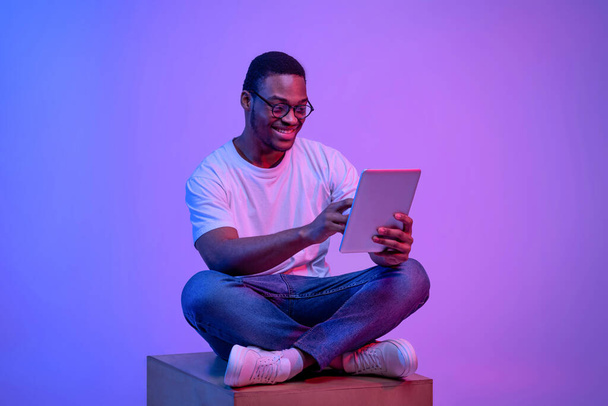 Молодий чорний фрілансер хлопець сидить з цифровим планшетом під яскравим неоновим світлом
 - Фото, зображення
