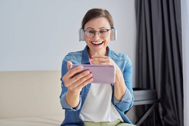 Reife positiv sprechende Frau mit Kopfhörer, die auf den Smartphone-Bildschirm schaut, Videoanruf - Foto, Bild