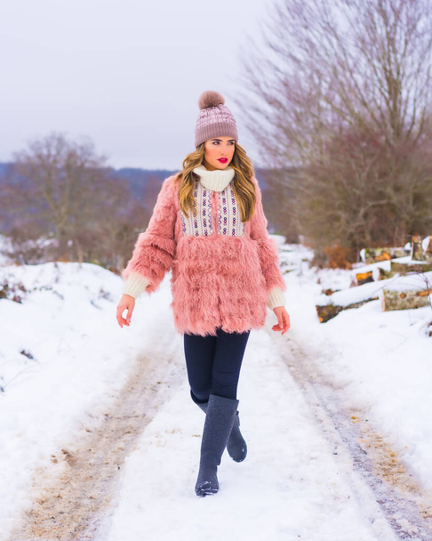 Young blonde girl with pink fur jacket and purple hat in the snow. Walking on a snow filled path, winter lifestyle - Foto, immagini