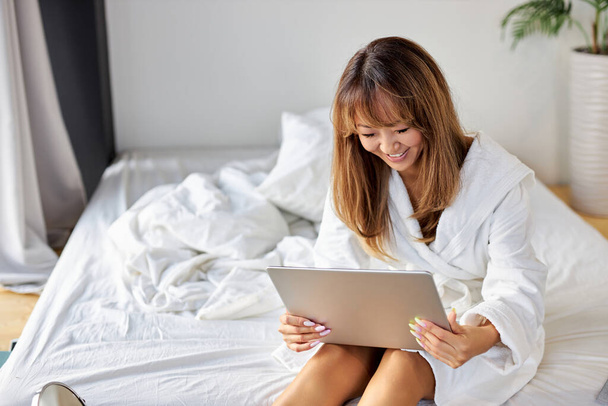 positive mixed race woman sitting on wide white bed using modern laptop - Фото, зображення