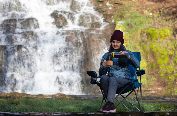 女性は滝の近くのキャンプチェアに座って、魔法瓶からお茶やコーヒーを飲む.  - 写真・画像