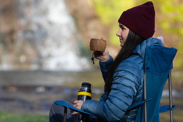 女性は滝の近くのキャンプチェアに座って、木製のカップKuksaからお茶やコーヒーを飲む.  - 写真・画像
