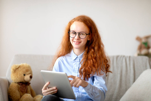 Dziewczyna w okularach za pomocą tabletu cyfrowego, siedzi na kanapie - Zdjęcie, obraz