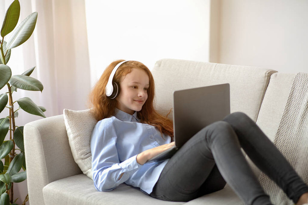 Девушка в наушниках отдыхает на диване и пользуется ноутбуком - Фото, изображение
