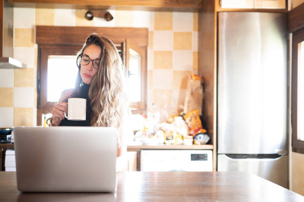 Mooie en elegante zakenvrouw voor haar laptop terwijl ze koffie drinkt in de keuken van haar huis. Nieuwe werkomstandigheden - Foto, afbeelding