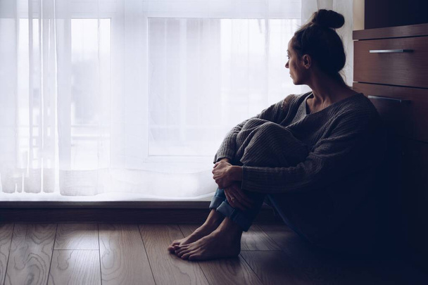Traurige Frau sitzt in ihrem Wohnzimmer auf dem Boden und schaut aus dem Fenster. - Foto, Bild