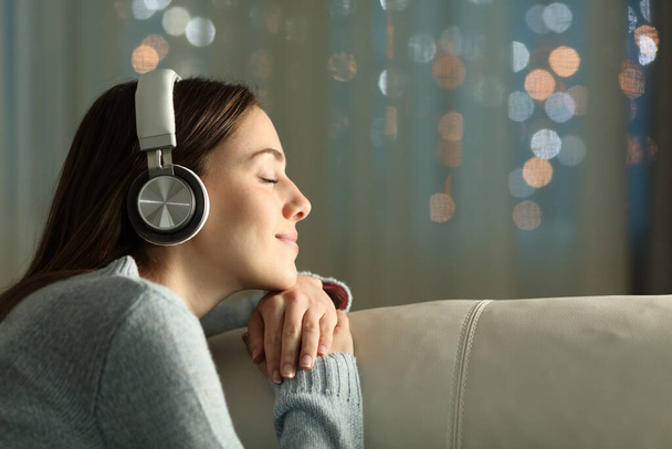 Profile of a woman with closed eyes feeling and listening to music with wireless headphones sitting on a couch in the night at home - Fotoğraf, Görsel