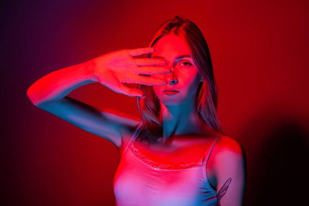 Unemotional young female covering eyes with hand in studio with red light - Foto, Imagem