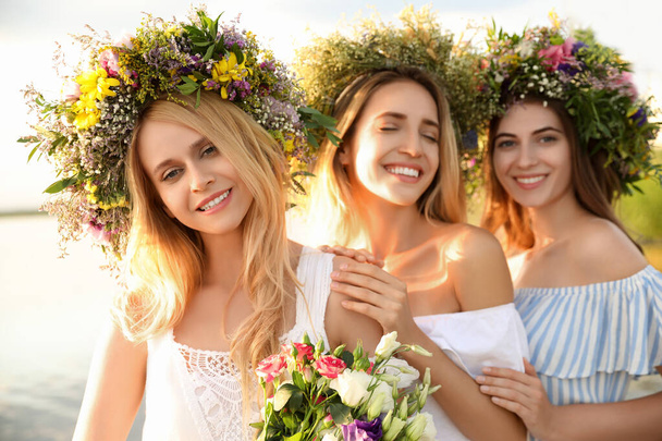 Young women wearing wreaths made of beautiful flowers near river on sunny day - Φωτογραφία, εικόνα