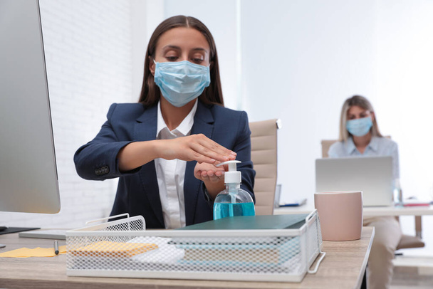 Office employee in mask applying hand sanitizer at workplace - Foto, afbeelding