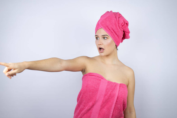 Giovane bella donna che indossa un asciugamano da doccia dopo il bagno in piedi su sfondo bianco isolato che punta con il dito sorpreso avanti, bocca aperta espressione stupita, qualcosa sul davanti - Foto, immagini