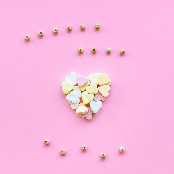Creative holiday concept of love. Heart made of colorful sugar candies on a pink background. Text Follow Your Heart made of wooden beads. Valentine's Day concept. Top view, flat lay. - Valokuva, kuva
