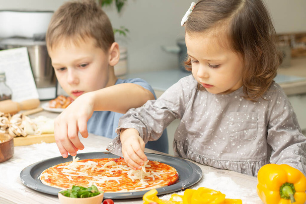 Aranyos kislány 2-4 szürke ruhában és fiú 7-10 pólóban pizzát főz együtt a konyhában. Testvérek főznek. - Fotó, kép