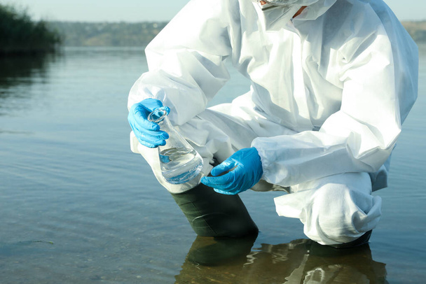 Scientist in chemical protective suit with conical flask taking sample from river for analysis, closeup - Foto, afbeelding
