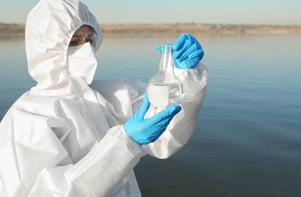 分析のために川からサンプルを取ってフローレンスフラスコと化学保護スーツの科学者 - 写真・画像
