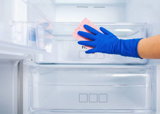 Το χέρι μιας γυναίκας σε ένα μπλε λαστιχένιο προστατευτικό γάντι και ένα ροζ σφουγγάρι πλένει και καθαρίζει τα ράφια του ψυγείου. Καθαριότητα, νοικοκυρά, δουλειές ρουτίνας. Κουτιά για λαχανικά, φρούτα και κρέας  - Φωτογραφία, εικόνα