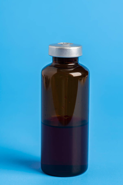 Ένα μακροσκοπικό πλάνο με επιλεκτική εστίαση ενός κεχριμπαρένιου χρώματος φιαλιδίου ένεσης εμβολίου σε ένα απλό μπλε φόντο. - Φωτογραφία, εικόνα