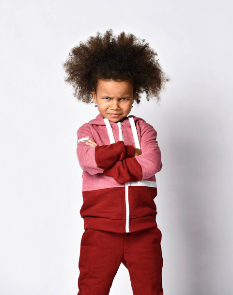 Sullen dark-skinned girl in a sports suit stands with folded arms on a gray background. - Fotografie, Obrázek