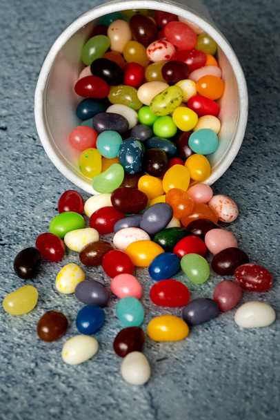  Jelly beans que se derrama de la taza de espuma blanca contra la mesa de fondo azul, composición festiva y colorida con candie, vista lateral con una sensación dinámica  - Foto, Imagen