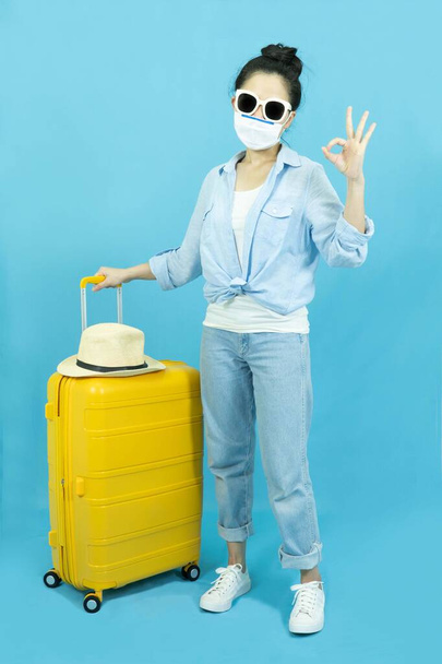Portrait of young beautiful asian woman wearing medical face mask in blue shirt with yellow suitcase or luggage isolated on blue background,Summer holidays, vacation and travel - Fotografie, Obrázek
