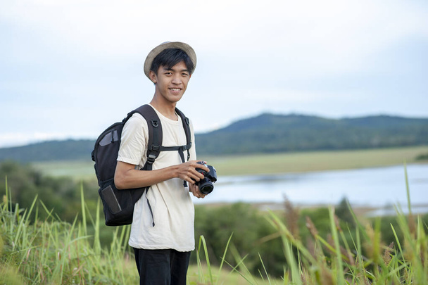 Mladý asijský muž cestovatel s batohem digitální fotoaparát pohled na fotoaparát s krásným přírodním jezerem pozadí. Koncept relaxační dovolené na cestách - Fotografie, Obrázek