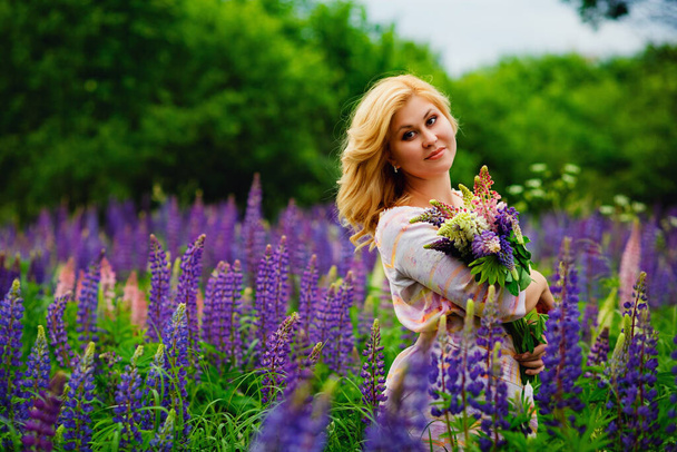 Beautiful young blonde on a green field among purple lupine wildflowers. Wildlife is a beautiful girl. - Photo, image