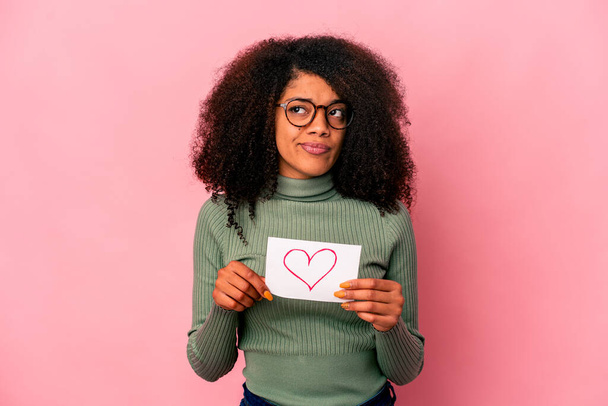 Νεαρή Αφροαμερικανή σγουρή γυναίκα που κρατά ένα σύμβολο καρδιάς σε πλακάτ μπερδεμένο, αισθάνεται αμφίβολο και αβέβαιο. - Φωτογραφία, εικόνα