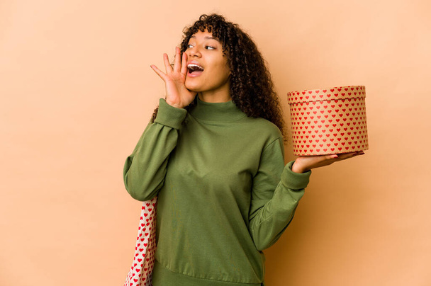 Joven afroamericana afro mujer sosteniendo un regalo de San Valentín gritando y sosteniendo la palma cerca de la boca abierta. - Foto, Imagen