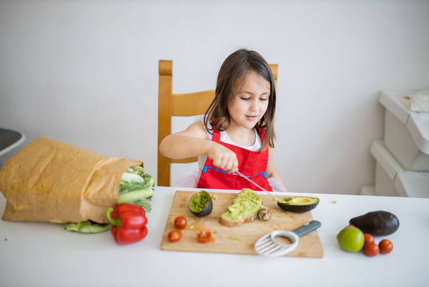klein meisje aan een tafel verspreiden avocado op een sneetje brood - Foto, afbeelding
