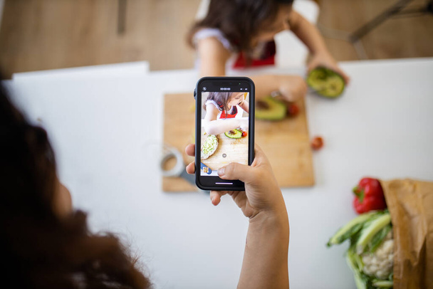 Mão feminina com um smartphone gravando uma menina cortando um abacate - Foto, Imagem
