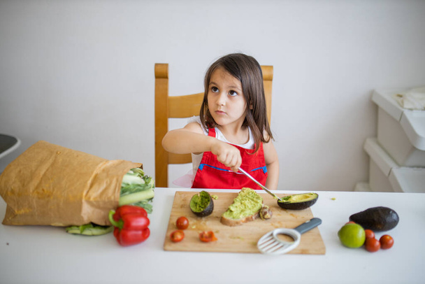 Очаровательная маленькая девочка за белым столом режет овощи на разделочной доске - Фото, изображение