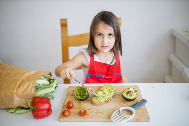 Маленькая девочка за столом, разбрасывающая авокадо на ломтик хлеба - Фото, изображение