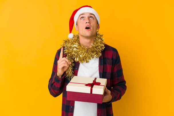 Nuori valkoihoinen mies, jolla on jouluhattu ja joka pitelee keltaisella pohjalla olevaa lahjaa ja osoittaa ylöspäin suu auki.. - Valokuva, kuva
