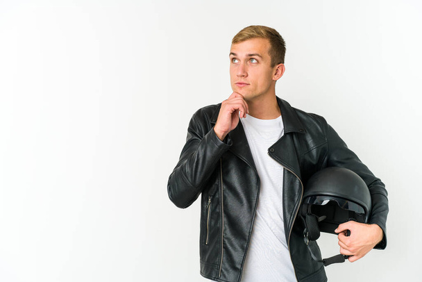 Jeune homme caucasien tenant un casque de moto isolé sur fond blanc regardant latéralement avec une expression douteuse et sceptique. - Photo, image
