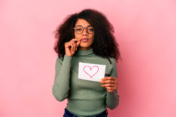 Νεαρή Αφροαμερικανή σγουρή γυναίκα που κρατά ένα σύμβολο καρδιάς σε πλακάτ με δάχτυλα στα χείλη κρατώντας ένα μυστικό. - Φωτογραφία, εικόνα