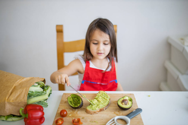 Little girl at a table spreading avocado on a slice of bread - Photo, Image