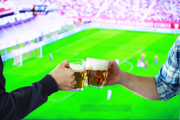 Hands horizontal holding lager beer glass and clinking on background of football game. Sport fans cheer up. Friends leisure lifestyle concept. - Foto, Bild