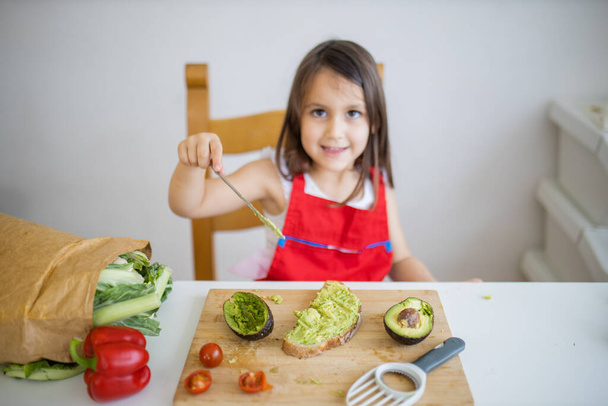Маленькая девочка за столом, разбрасывающая авокадо на ломтик хлеба - Фото, изображение