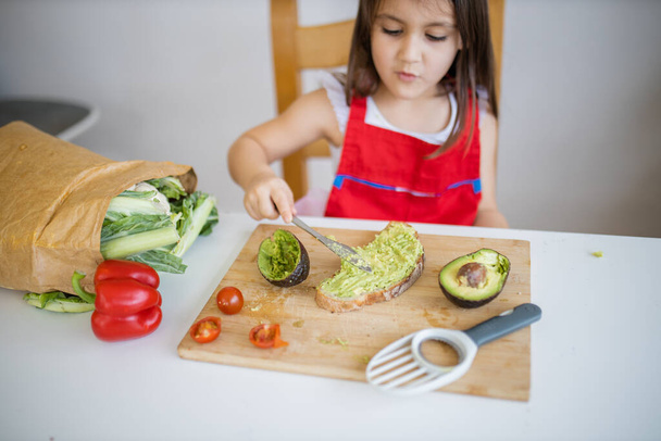 klein meisje aan een tafel verspreiden avocado op een sneetje brood - Foto, afbeelding