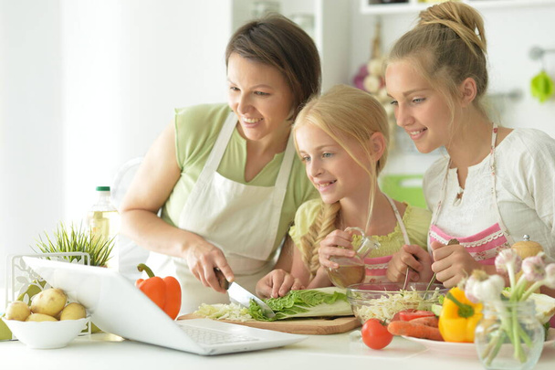 Leuke meisjes met moeder koken en kijken naar laptop in de keuken - Foto, afbeelding