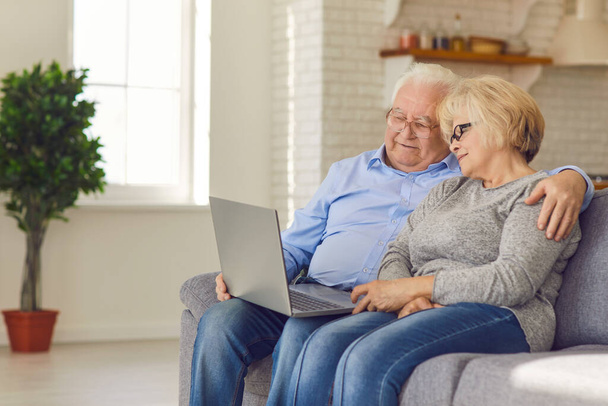 Loving happy elderly couple sitting together on sofa, hugging and watching movie online - Foto, imagen