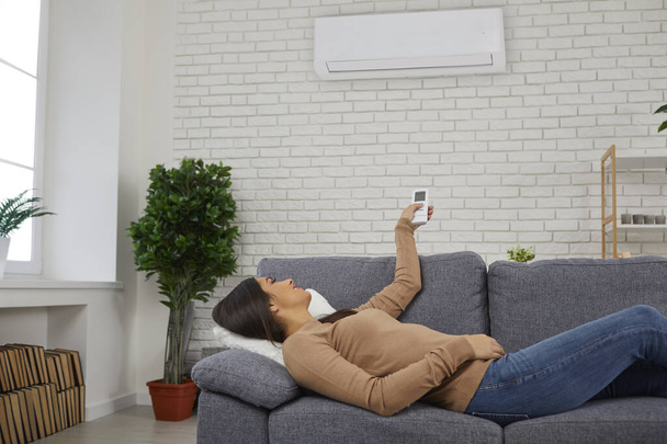 Woman lying on couch at home and switching on air conditioner with remote control - Fotografie, Obrázek