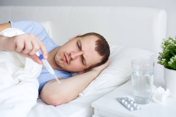 egészségügy és betegség koncepció - portré beteg ember láz fekszik az ágyon, és nézi a hőmérő - Fotó, kép