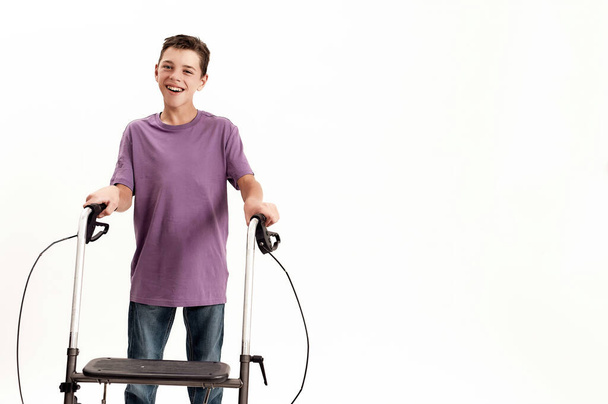 Happy teenaged disabled boy with cerebral palsy smiling at camera, taking steps using his walker isolated over white background - 写真・画像
