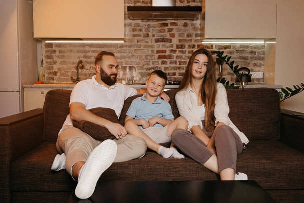 Dad, son, and a young mom with long hair are watching with interest TV on the sofa in the apartment. The family is enjoying a happy evening at home. The father is staring at his boy and wife. - Photo, Image