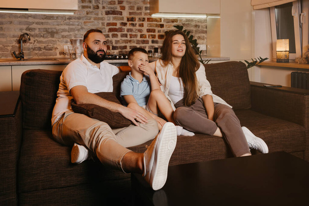 Dad with a beard, son, and young mom with long hair are watching TV and smiling on the sofa. The family is enjoying a happy evening at home. A child is laughing and covering his mouth with his hand. - Photo, Image