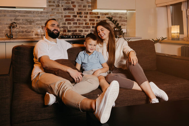 Тато з бородою, сином і молодою мамою з довгим волоссям дивиться телевізор і посміхається на диван. Сім'я насолоджується щасливим вечором вдома. Мати сміється
 - Фото, зображення